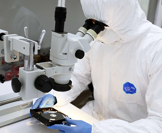 Technician in cleanroom attire examining a hard drive under a microscope.